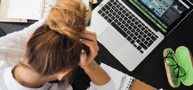 Top-down image of a woman sitting at a cluttered desk. She's holding her head and looking down.